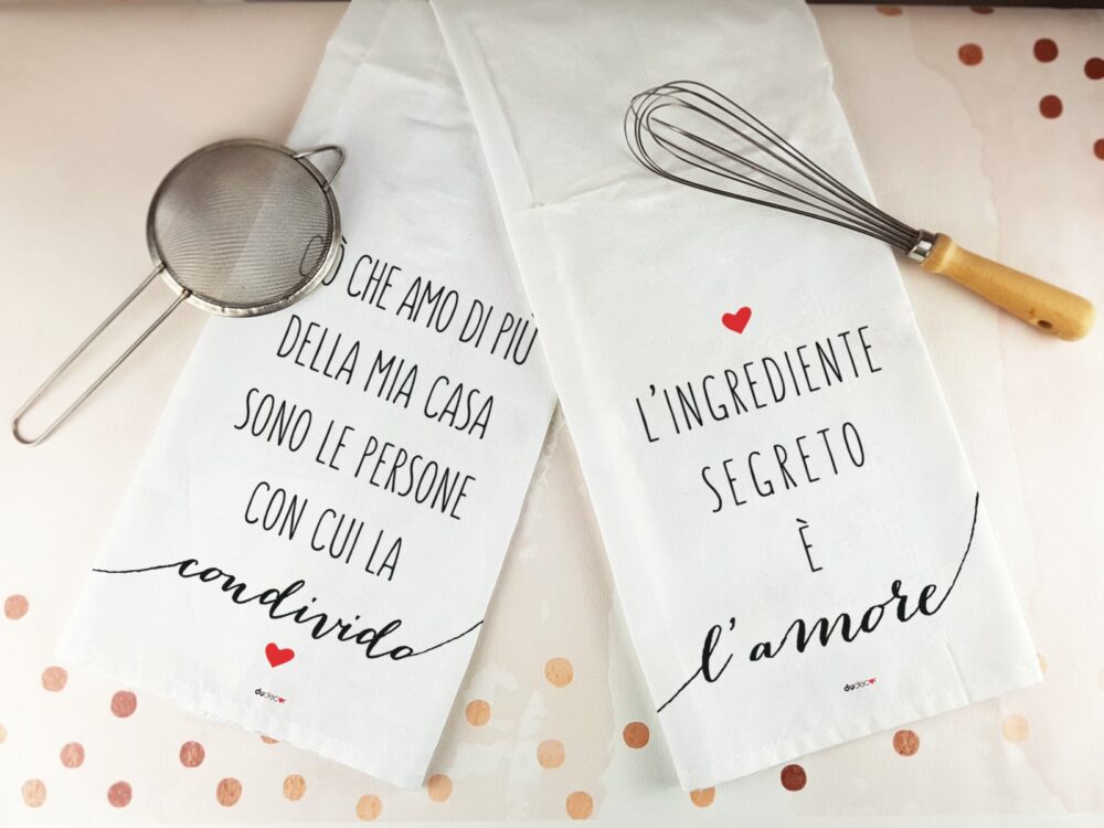 Canovacci da cucina con frasi Dudecor appoggiati sul piano di lavoro insieme a utensili