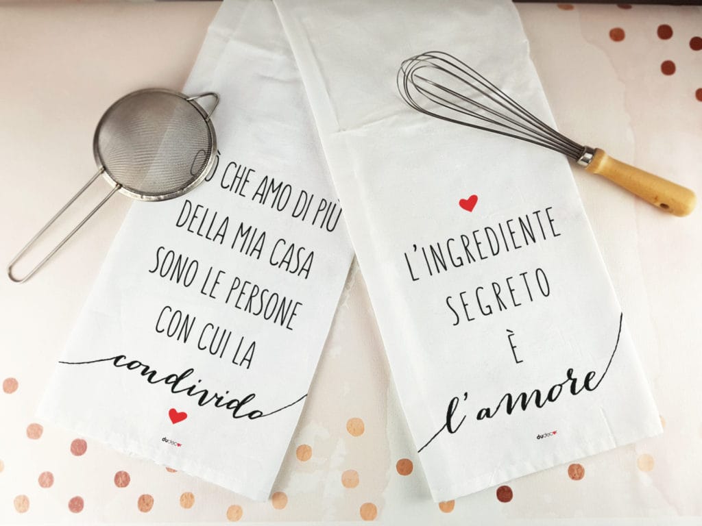 Canovacci da cucina con frasi Dudecor appoggiati sul piano di lavoro insieme a utensili