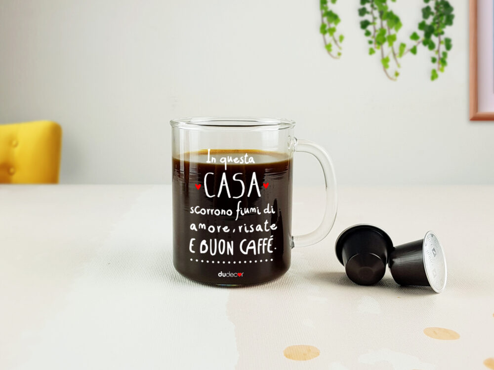 Tazza in vetro Dudecor con scritta “In questa casa scorrono fiumi di amore, risate e buon caffè” appoggiata su tavolo chiaro con caffè appena fatto