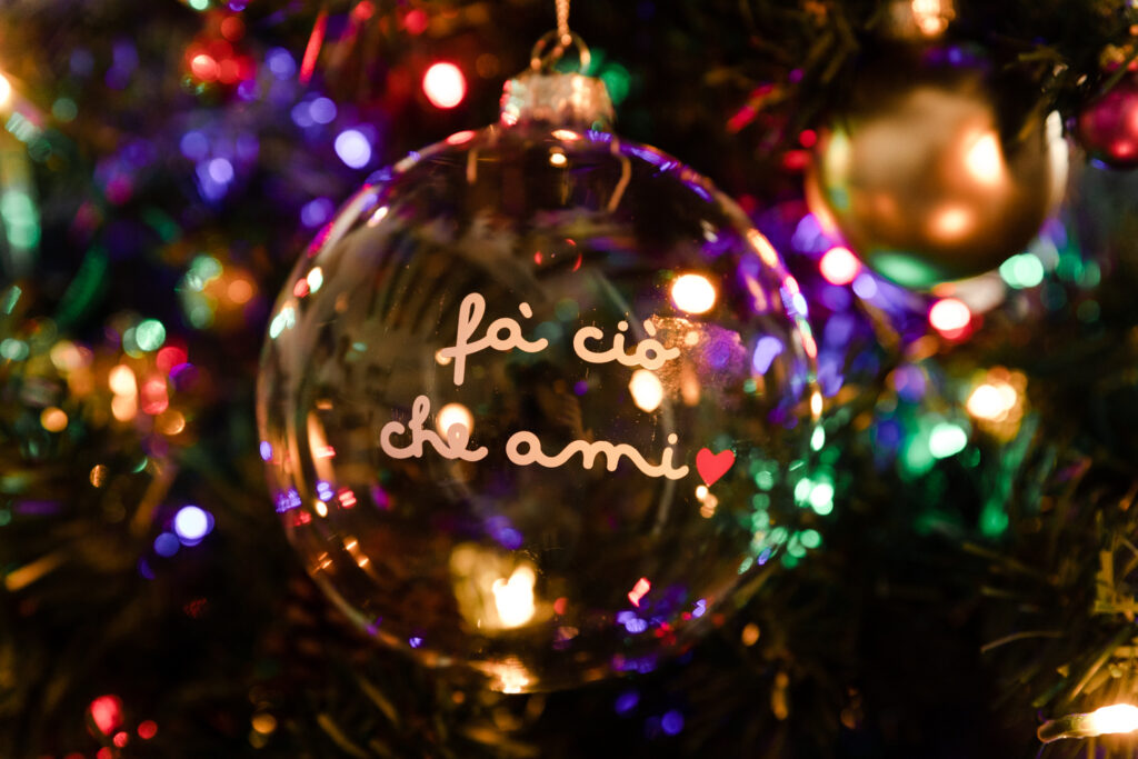 Pallina natalizia trasparente Dudecor con la scritta “fai ciò che ami” e un cuore rosso, appesa all’albero e illuminata da luci colorate.