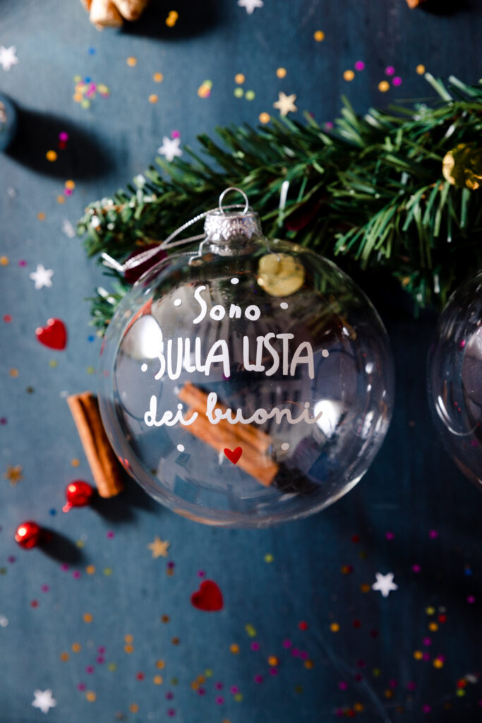 Pallina natalizia trasparente Dudecor con la scritta “Sono sulla lista dei buoni”, appoggiata su sfondo blu con decori, cannella e rami verdi.