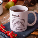 Tazza bianca Dudecor con la definizione di “Natale”, riempita di cioccolata calda, appoggiata su un tavolo in legno con cannella, frutta e luci natalizie.