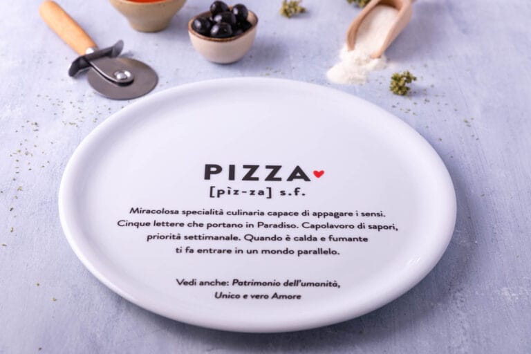 Piatto in ceramica bianca con definizione ironica della parola pizza, fotografato con ingredienti e utensili da cucina sullo sfondo.