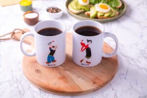 Coppia di tazze in ceramica con illustrazioni stilizzate di personaggi ispirati a Popeye e Olivia, appoggiate su tagliere in legno con colazione sullo sfondo.