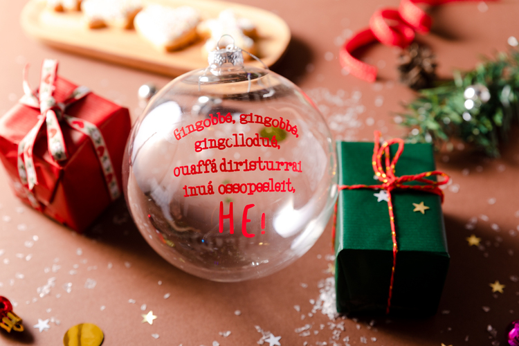 Pallina di Natale Dudecor con la frase ironica che richiama “Jingle Bells” cantata in modo storpiato, accanto a pacchetti regalo e decorazioni festive.