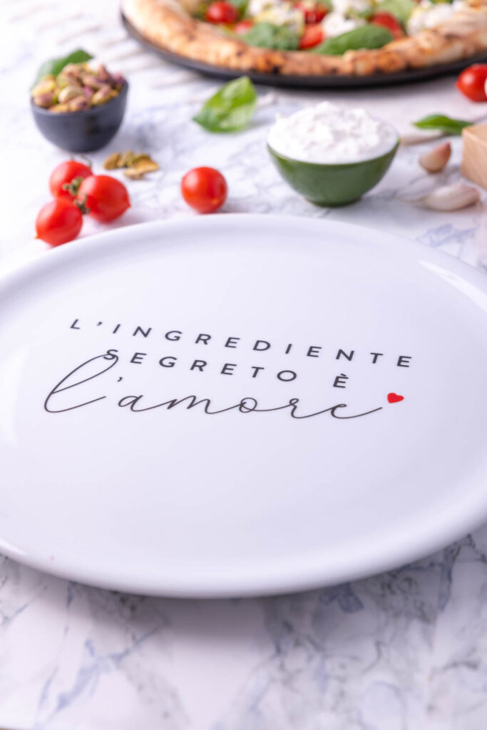 Piatto in ceramica Dudecor con scritta “L’ingrediente segreto è l’amore” ambientato su tavola con ingredienti per pizza