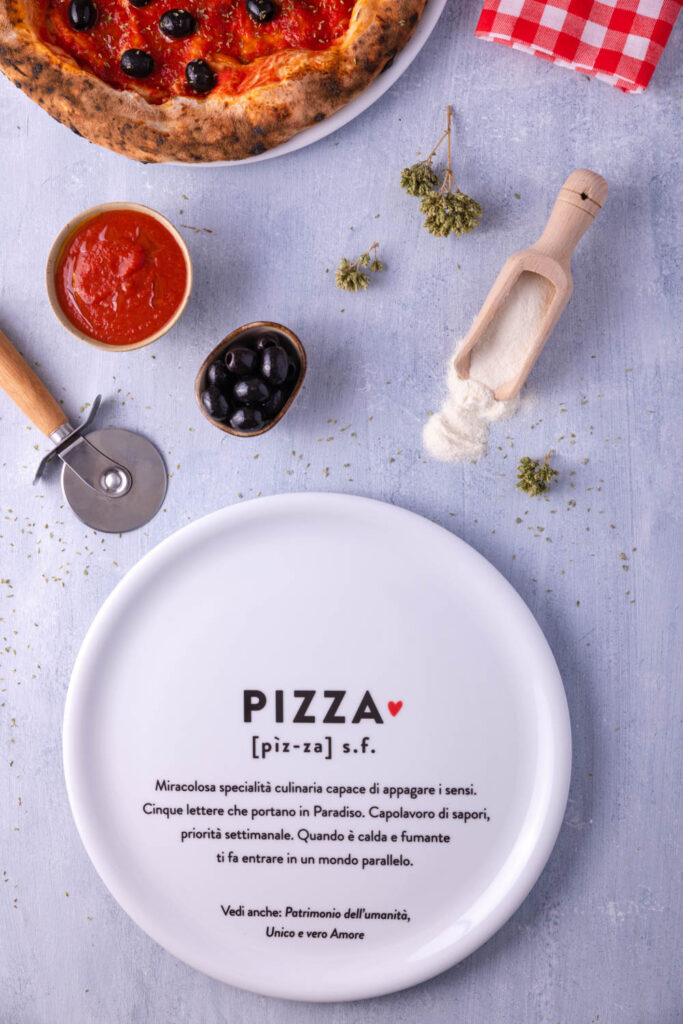 Piatto pizza in ceramica con definizione ironica della parola pizza, apparecchiato con ingredienti e accessori su tavola chiara