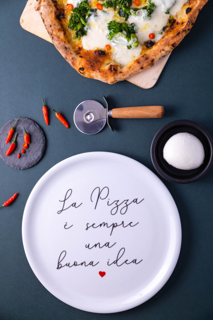 Piatto pizza in ceramica bianca con scritta “La pizza è sempre una buona idea”, apparecchiato con pizza, taglia pizza e ingredienti su tavola scura